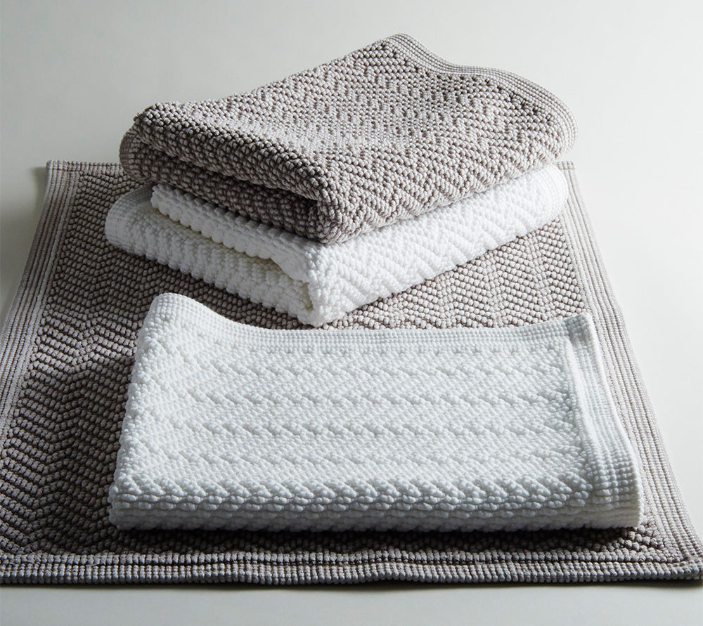 Hochwertiger Badvorleger ZIG ZAG in gestapelter Ansicht, mit charakteristischem Zickzack-Muster in Weiss und Grau. Moderne Baumwollvorleger in verschiedenen Groessen zeigen die elegante Struktur und Textur. Badvorleger ZIG ZAG Schwarz, Hersteller: Evelyn Kahle.