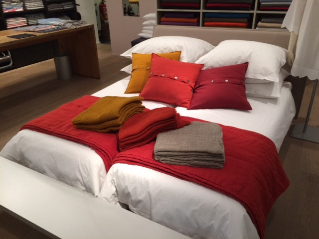 Bettwäsche Bianco Gewaschene Baumwolle: Stilvolles Bett mit weißer Bettwäsche, rotem Überwurf und dekorativen Kissen in Rot und Senfgelb, präsentiert in einem modernen Showroom mit braunem Holzboden und Stoffregalen im Hintergrund. Hochwertige Texturen und Farben zeigen die Premium-Qualität der gewaschenen Baumwolle.