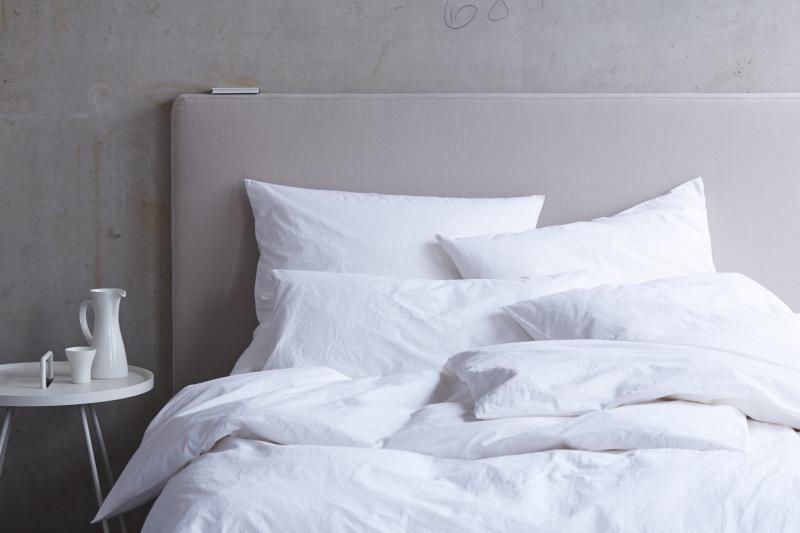 Bettwäsche Bianco Gewaschene Baumwolle: Luxuriöse weisse Bettwäsche aus vorgewaschener Baumwolle auf grauem Bett, lässig drapiert mit mehreren Kissen, daneben weisser Beistelltisch mit Kanne. Minimalistisches, helles Design für modernen Schlafkomfort.