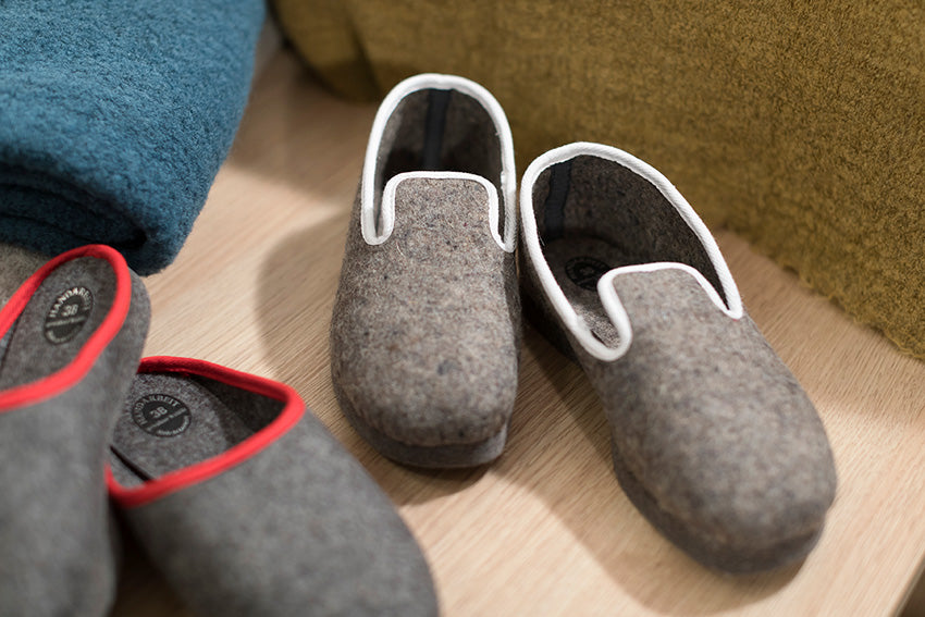 Geschlossene graue Wollfilz-Hausschuhe mit weissem Einfassband, handgefertigt in Deutschland, auf Holzboden neben anderen Pantoffeln fotografiert. Hausschuhe geschlossen, Band weiss - Pantoffeln, Hersteller: Stoffkontor