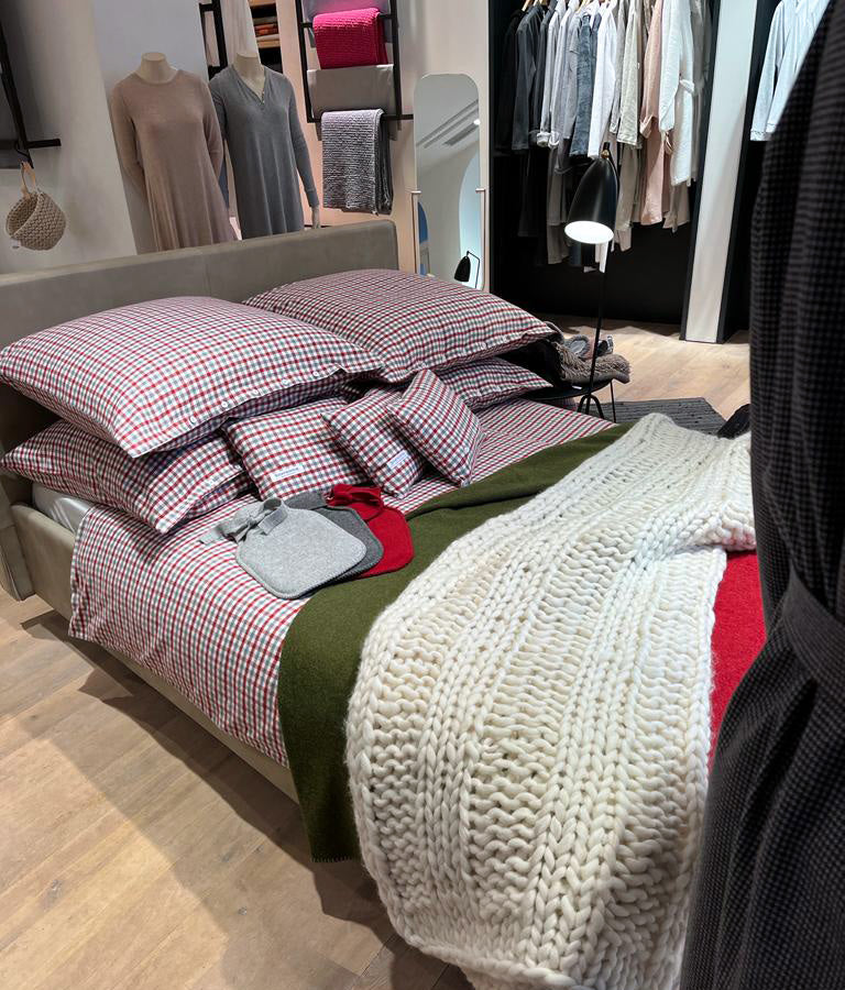 Bettwäsche Klassisches Karo Flanell Rot: Hochwertige Bettwäsche aus Portugal mit klassischem rot-weißem Karomuster, präsentiert auf einem Ausstellungsbett mit gestrickter weißer Decke und grünem Akzent. Stoffkontor-Qualität mit praktischer Knopfleiste, aus reiner Baumwolle gefertigt.