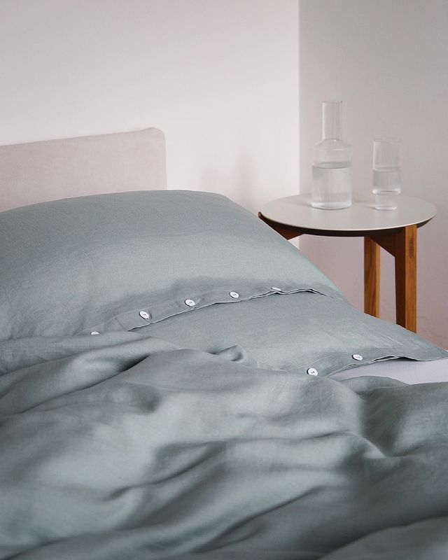 Elegante Bettwäsche aus vorgewaschenem Leinen in sanftem Salbeigrün, mit klassischer Knopfleiste. Stilvolle Produktaufnahme in einem Schlafzimmer zeigt die natürliche Textur der Bettwäsche neben einem minimalistischen Nachttisch mit Glaskaraffen.