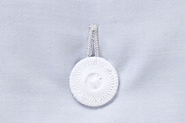 Eleganter weißer Wäscheknopf der Bettwäsche Perla Damast, mit dekorativer Prägung auf hellblauem Satinstoff. Hochwertige Verarbeitung mit gezwirnter Knopfleiste zeigt handwerkliche Qualität und stilvolles Design.