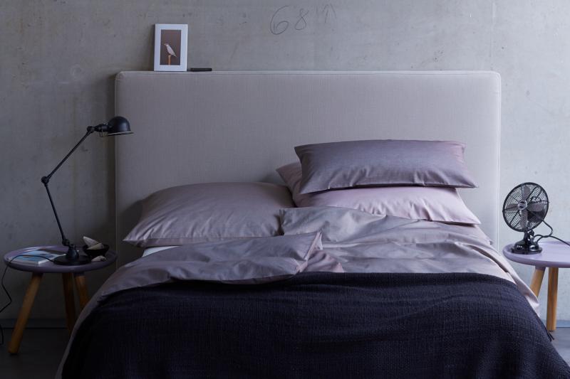 Bettwäsche Potassio Damast in elegantem Rosenholzton auf stilvollem Bett mit grauer Kopfstütze, dunkler Tagesdecke und minimalistischen Nachttischen. Stimmungsvolle Präsentation der hochwertigen Damast-Bettwäsche mit Vintage-Tischventilator und Leselampe als Accessoires.