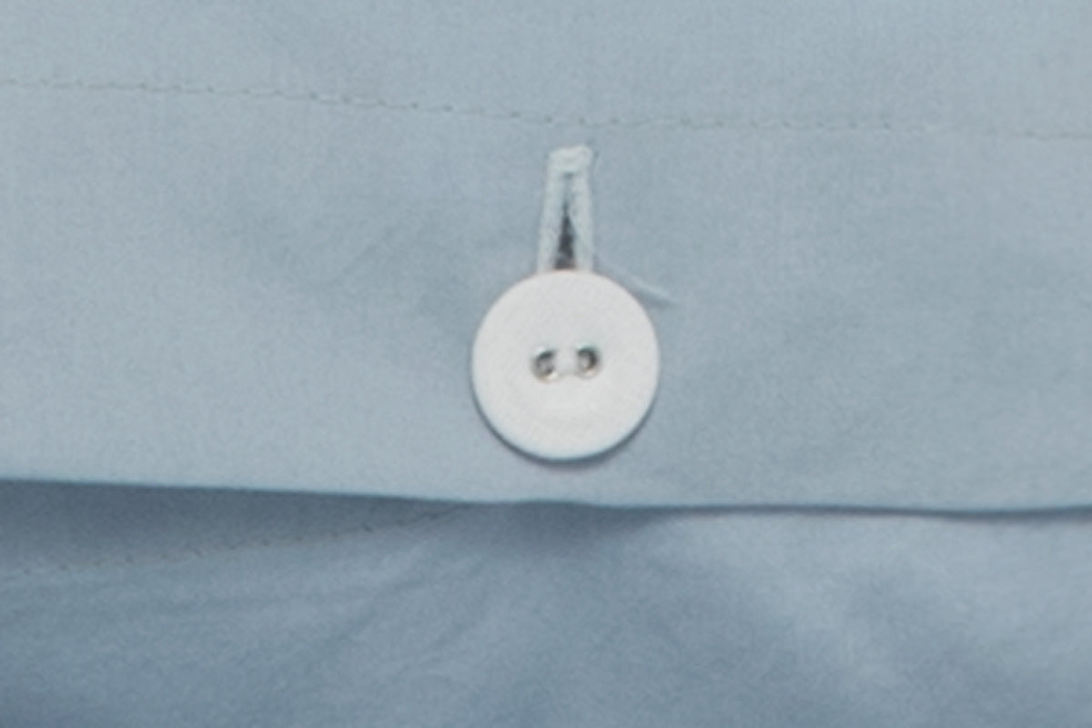 Nahaufnahme eines weissen Stoffknopfes auf hellblauer Bettwäsche Lagoon aus gewaschener Baumwolle, Detail der hochwertigen Verarbeitung mit Knopfschlaufe, harmonische Farbkombination für zeitloses Schlafzimmerdesign.