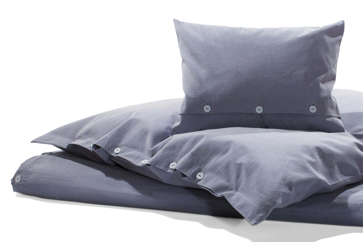 Bettwäsche Stonewashed Denim Ciclamino: Hochwertige Bettwäsche in softem Lavendelblau-Grau, stonewashed verarbeitet mit Knopfverschluss. Die Baumwollbettwäsche im Jeans-Look liegt lässig auf einem weißen Bett, zeigt Kissen und Bettdecke in harmonischer Anordnung.