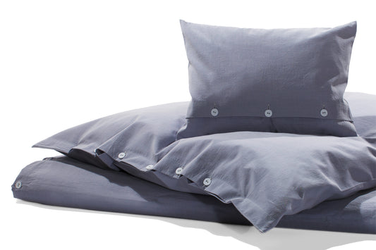 Bettwäsche Stonewashed Denim Ciclamino: Hochwertige Bettwäsche in softem Lavendelblau-Grau, stonewashed verarbeitet mit Knopfverschluss. Die Baumwollbettwäsche im Jeans-Look liegt lässig auf einem weißen Bett, zeigt Kissen und Bettdecke in harmonischer Anordnung.