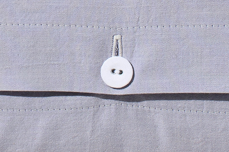Detailaufnahme der Bettwäsche Stonewashed Denim Ciclamino: Weißer Stoffknopf auf grau-lavendelfarbenem Baumwollstoff mit feiner Naht, charakteristisch für die hochwertige Stonewashed-Verarbeitung. Zeigt die handwerkliche Qualität der italienischen Webkunst.