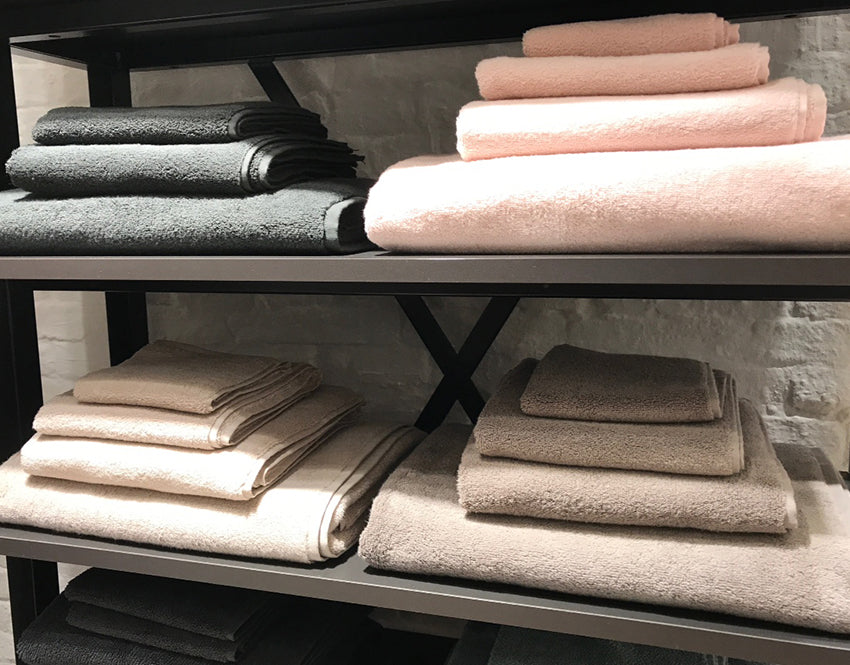 Luxuriöses Handtuch Dreampure Weseta in Stone Grey, sorgfältig gefaltet auf einem dunklen Regal präsentiert, zusammen mit weiteren hochwertigen Handtüchern in verschiedenen Größen und sanften Pastelltönen. Handtücher, Hersteller: Weseta Switzerland